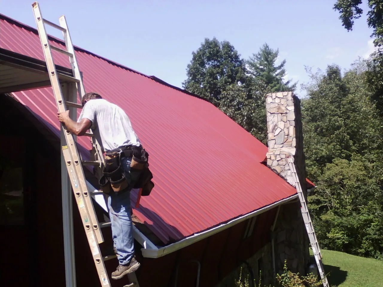 Roofer on a ladder beside a red metal roof and stone chimney in Niota